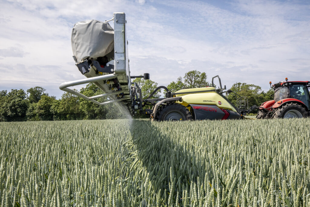 The photo shows a modern agricultural scene where a tractor is pulling a large piece of farming equipment through a field of crops, likely wheat. The equipment appears to be a sprayer, specifically an AEON Centura Line machine, applying liquid evenly across the field. The sprayer's boom extends horizontally over the crops, with mist being emitted from nozzles, indicating the application of pesticides or fertilizers. The scene is set under a partly cloudy sky with a background of dense green trees.